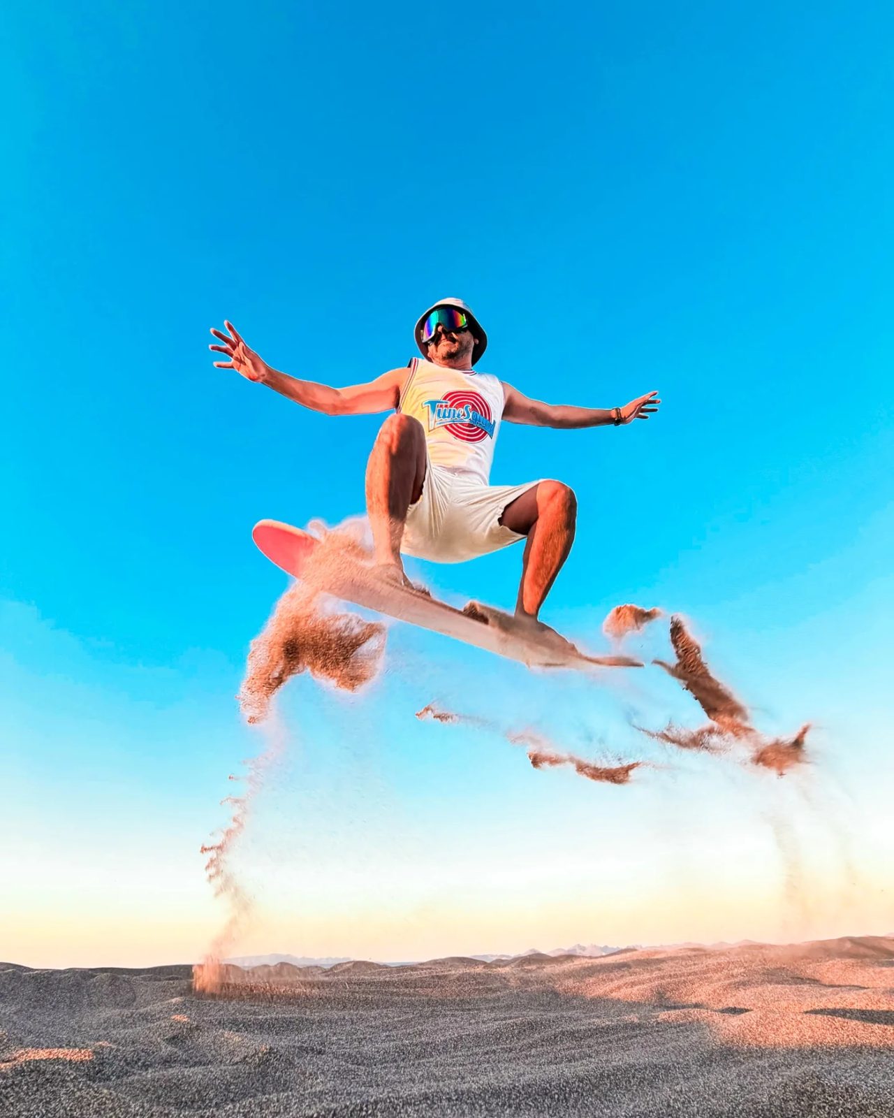 Sandboarder saltando en el aire con la tabla y arena volando sobre las dunas de El Mogote y el cielo azul
